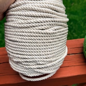 May include: A tightly wound coil of off-white rope. The rope is thick and appears to be made of twisted fibers. The coil is held by a hand, and the background is a blurred view of a green lawn and a red wooden surface.