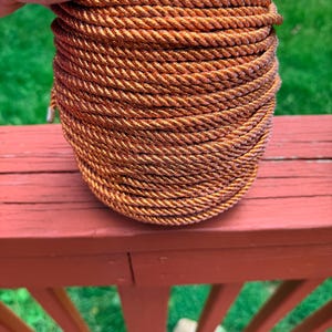 May include: A spool of copper-colored rope. The rope is tightly wound and has a textured appearance. The spool is resting on a red wooden surface with a green grassy background.