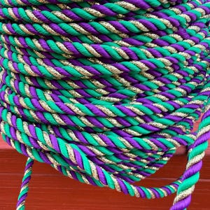 May include: A tightly coiled rope in a vibrant color scheme of green, purple, and gold. The rope is made of twisted strands, creating a textured surface. The gold strands add a touch of shimmer. The rope is likely used for decorative purposes.