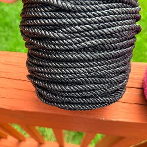 May include: A tightly wound coil of black rope. The rope is made of a thick, textured material and is wound in neat, even layers. The rope is held by a hand, and the background is a blurred view of green grass and a red wooden surface.