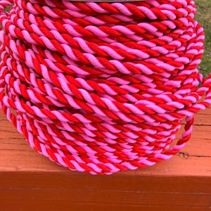 May include: A spool of twisted rope in alternating shades of red and pink. The rope is tightly wound, creating a textured surface. The spool sits on a wooden surface, with a white cardboard top.