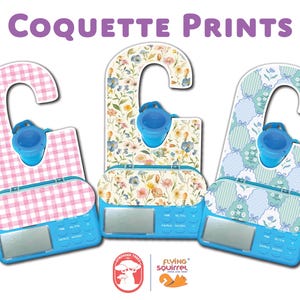 May include: Three blue and white baby scales with decorative patterned tops. The patterns include pink gingham, floral, and blue and white bows. The scales have a digital display and buttons. The text "Coquette Prints" is at the top.
