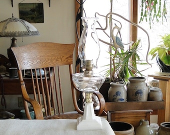 May include: A clear glass oil lamp with a white base sits on a white tablecloth. The lamp has a brass-colored burner and a tall, clear glass chimney. The background includes a wooden chair, pottery, and a window.