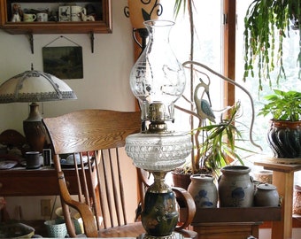 May include: A vintage oil lamp with a clear glass shade and a decorative base. The lamp is sitting on a table with a white tablecloth. There are other decorative items on the table, including a vase, a pitcher, and a bowl.