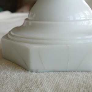 May include: Close-up of a white, opaque, pedestal base with a hexagonal shape. The base has a smooth, glossy finish with subtle variations in the surface. The base is sitting on a white, textured surface.