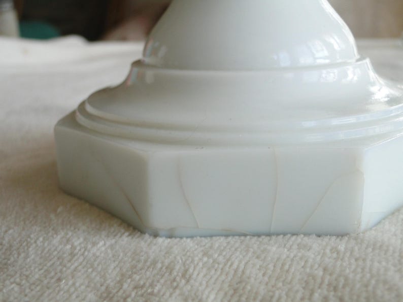 May include: Close-up of a white, opaque, pedestal base with a hexagonal shape. The base has a smooth, glossy finish with subtle variations in the surface. The base is sitting on a white, textured surface.