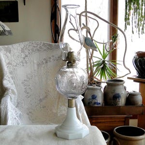 May include: A clear glass oil lamp with a decorative globe and a white base. The lamp has a brass-colored collar and a clear glass chimney. The base is hexagonal. The lamp is on a white surface.