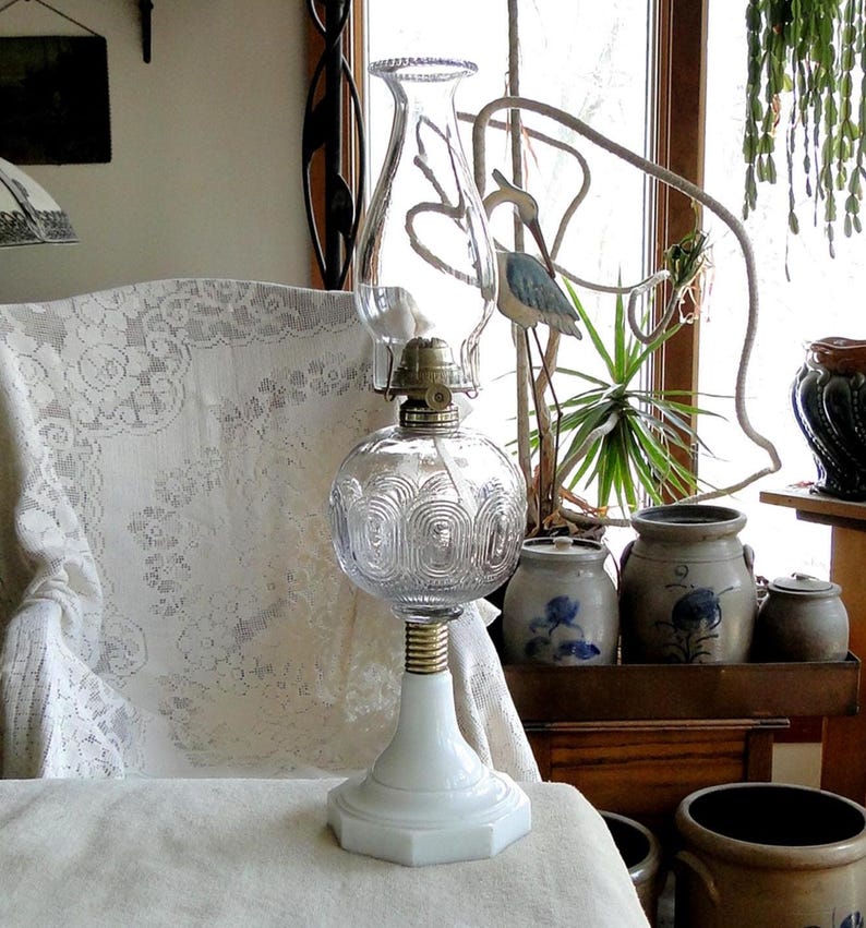 May include: A clear glass oil lamp with a decorative globe and a white base. The lamp has a brass-colored collar and a clear glass chimney. The base is hexagonal. The lamp is on a white surface.