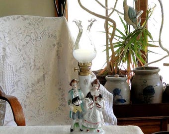 May include: A vintage porcelain oil lamp featuring a couple in period clothing. The lamp has a clear glass chimney and a brass base. The figures are dressed in white, black, and light blue, with a touch of red. The lamp is on a white surface.