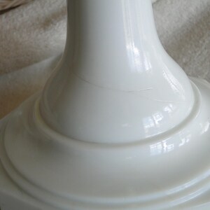 May include: Close-up of a white, glossy, ceramic base with a flared, rounded top and a square, stepped bottom. The base has a smooth, reflective surface with a subtle crack. The design is simple and elegant.