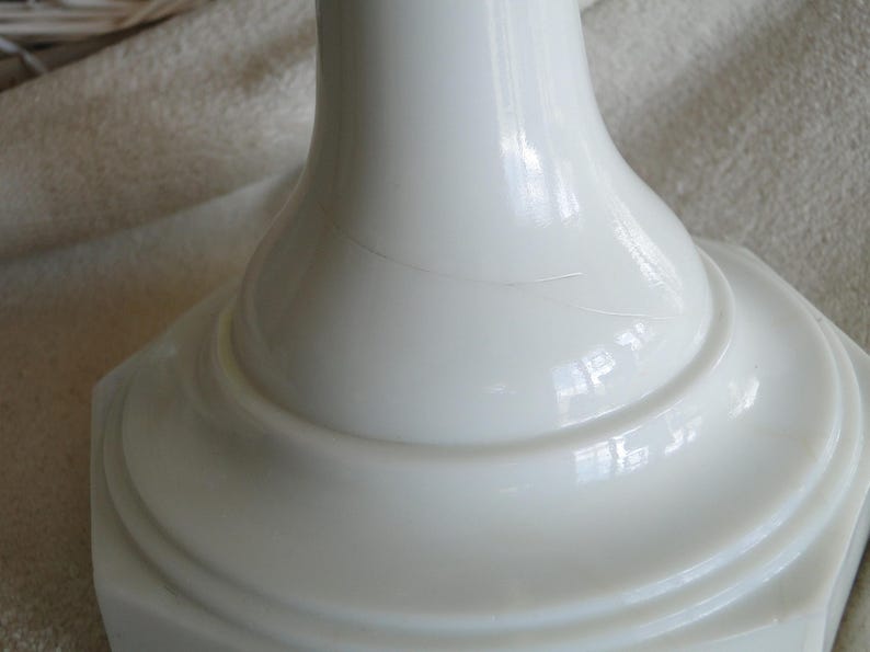 May include: Close-up of a white, glossy, ceramic base with a flared, rounded top and a square, stepped bottom. The base has a smooth, reflective surface with a subtle crack. The design is simple and elegant.