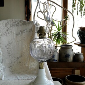 May include: A vintage oil lamp with a clear glass globe and chimney. The lamp has a white base and a brass-colored connector. The globe features a decorative pattern. The lamp is displayed on a white surface.