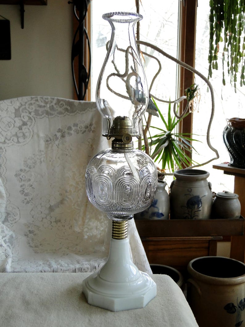 May include: A vintage oil lamp with a clear glass globe and chimney. The lamp has a white base and a brass-colored connector. The globe features a decorative pattern. The lamp is displayed on a white surface.