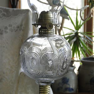 May include: Clear glass oil lamp with a round, patterned reservoir and a brass base. The lamp has a glass chimney and a metal top. The lamp is a vintage style and is likely used for home decor.