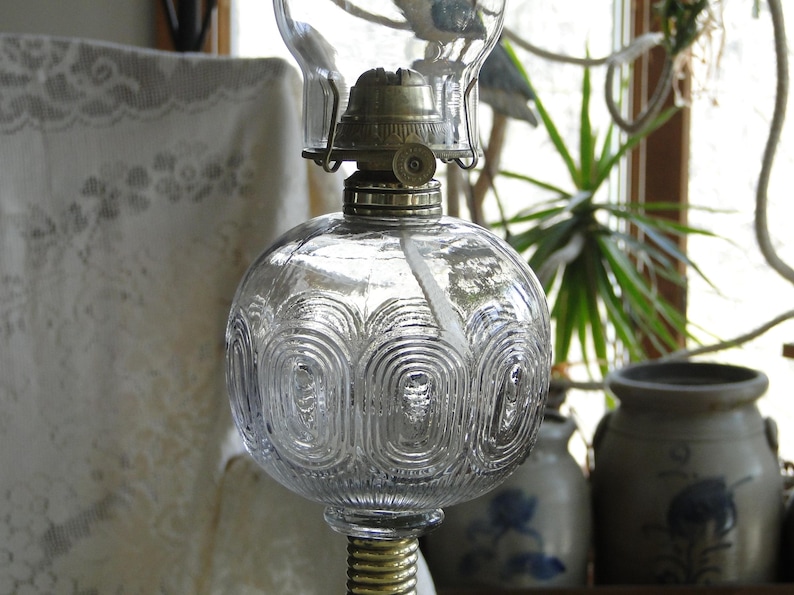 May include: Clear glass oil lamp with a round, patterned reservoir and a brass base. The lamp has a glass chimney and a metal top. The lamp is a vintage style and is likely used for home decor.