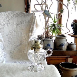 May include: A clear glass oil lamp with a brass top and a decorative base. The lamp is sitting on a white surface, with a lace-covered chair and various antique pottery pieces in the background. A decorative metal heron stands near a window.