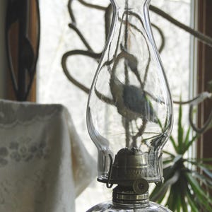 May include: A clear glass oil lamp with a decorative, textured base and a clear glass chimney. The lamp has a metal burner and a wick. The background includes a window and a lace tablecloth.