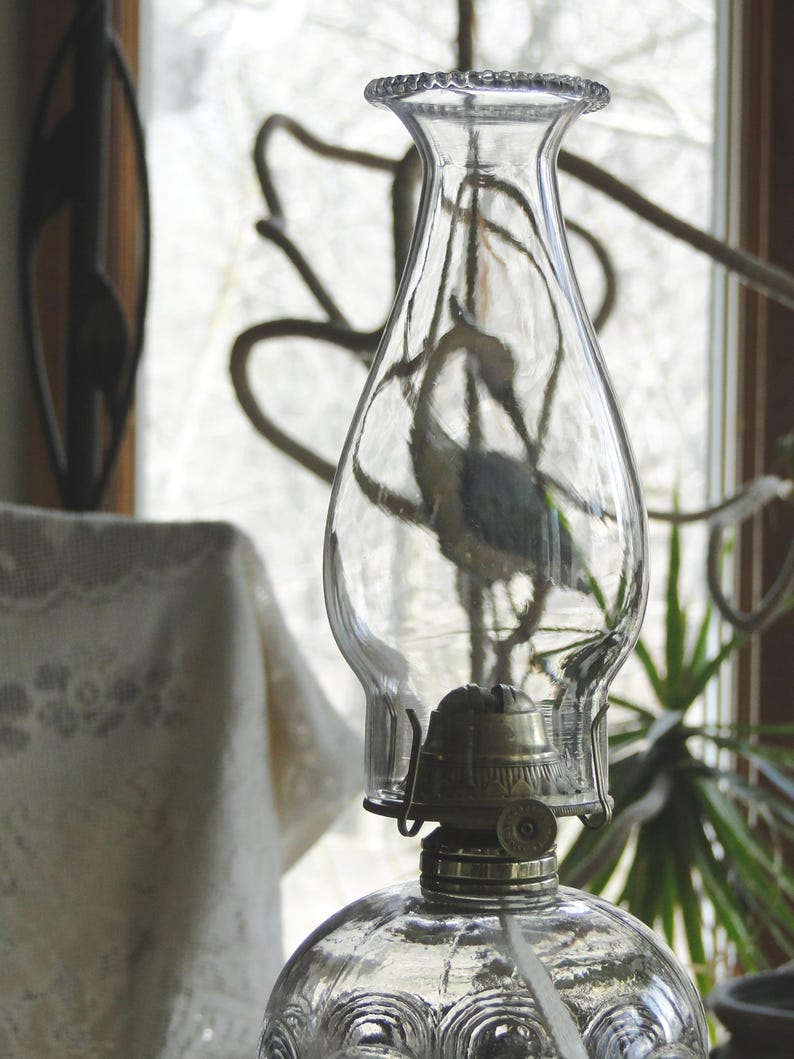 May include: A clear glass oil lamp with a decorative, textured base and a clear glass chimney. The lamp has a metal burner and a wick. The background includes a window and a lace tablecloth.