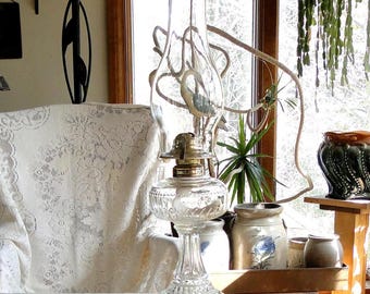 May include: A clear glass oil lamp featuring a textured, patterned base and a tall, bulbous chimney. The lamp has a metallic burner assembly situated between the glass reservoir and the chimney, with a transparent, reflective finish.