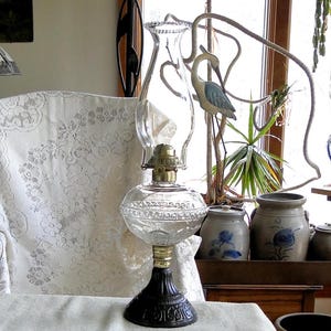 May include: A vintage oil lamp with a clear glass globe and a black base. The lamp has a brass burner and a decorative etched pattern around the globe. The lamp is sitting on a white surface, with a lace-covered chair in the background.