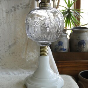 May include: A clear glass oil lamp with a round, textured globe and a white base. The lamp has a brass-colored metal top and a white, hexagonal base. The globe features a repeating pattern of curved lines. The lamp is designed for decorative or functional use.