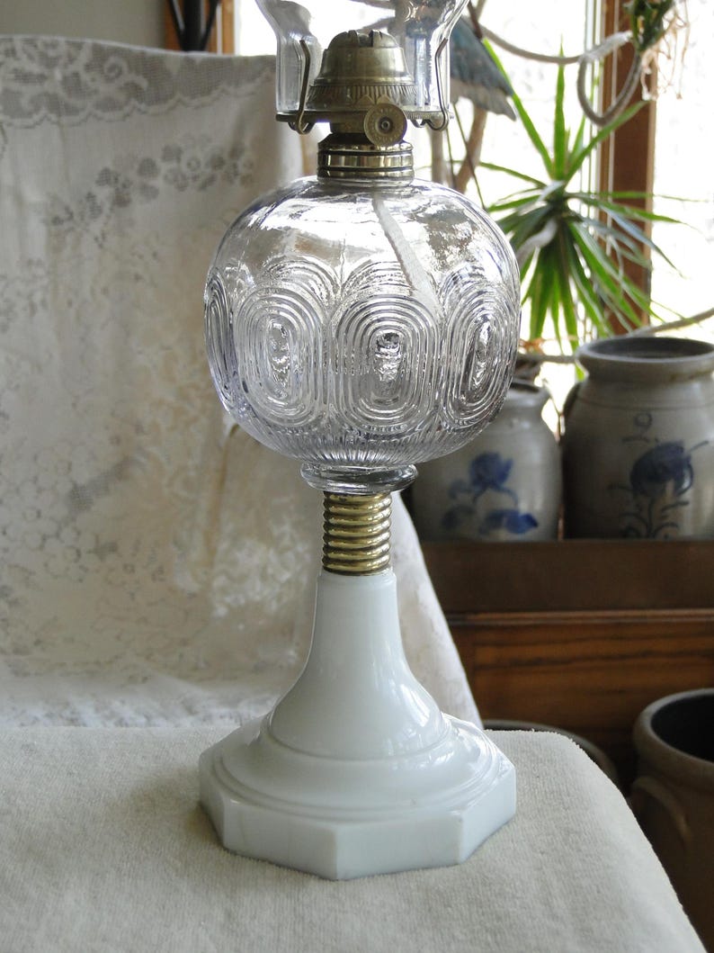 May include: A clear glass oil lamp with a round, textured globe and a white base. The lamp has a brass-colored metal top and a white, hexagonal base. The globe features a repeating pattern of curved lines. The lamp is designed for decorative or functional use.
