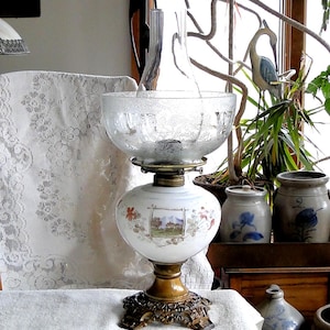 May include: A white oil lamp with a floral design and a clear glass chimney. The lamp is on a gold metal base with a decorative design. The lamp is sitting on a white tablecloth.