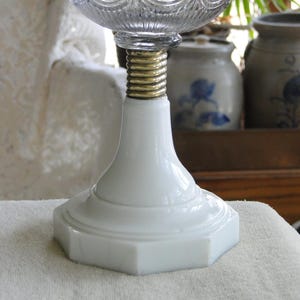 May include: A white, octagonal-based lamp with a clear, textured glass top. The lamp features a gold-colored, threaded metal section connecting the base to the glass. The lamp is likely a decorative or functional lighting fixture.