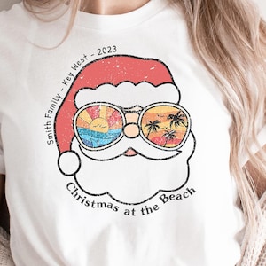 May include: White t-shirt featuring a Santa Claus graphic with sunglasses reflecting a beach scene. The design includes the text "Smith Family - Key West - 2023" and "Christmas at the Beach". The shirt has a vintage, distressed look.