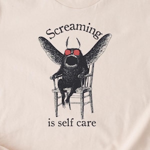 May include: A beige t-shirt with a black and white illustration of a creature with wings and sunglasses sitting on a chair. The text "Screaming is self care" is printed below the illustration.