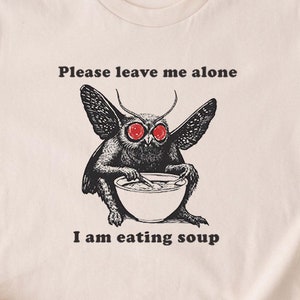 Peut inclure: Un t-shirt beige avec une illustration en noir et blanc d'un papillon de nuit aux yeux rouges en train de manger de la soupe dans un bol. Le texte sur le t-shirt dit "Veuillez me laisser tranquille, je suis en train de manger de la soupe."