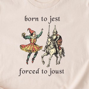 以下が含まれることがあります： ベージュ色のTシャツ。中世風のイラストで、道化師と馬に乗った騎士が描かれています。「born to jest forced to joust」の文字が図の上と下にプリントされています。柔らかい素材のシャツです。