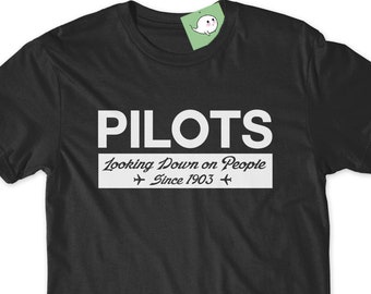 以下が含まれることがあります： 白い文字で「PILOTS Looking Down on People Since 1903」と書かれた黒いTシャツ。テキストの両側に小さな飛行機のグラフィックがあります。