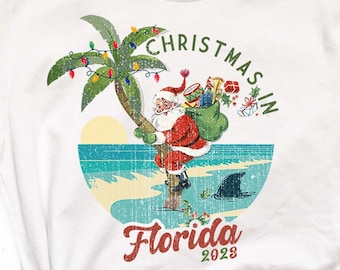 Może przedstawiać: Biała koszulka z grafiką Świętego Mikołaja na plaży na Florydzie, z palmą ozdobioną lampkami choinkowymi. Tekst brzmi "Christmas in Florida 2023". Koszulka ma zawiązany węzeł na dole.