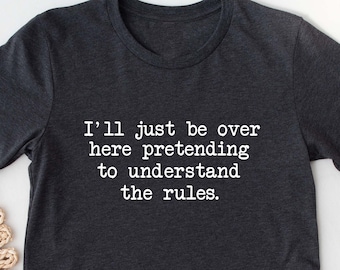 May include: A dark gray t-shirt with the text "I'll just be over here pretending to understand the rules." printed in white.