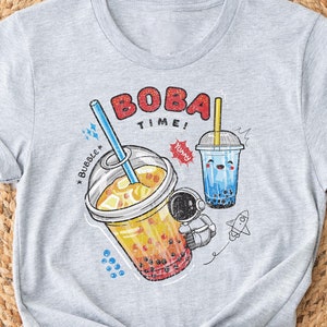 May include: A light grey t-shirt with a graphic of two boba tea drinks, one orange and one blue, and the words "BOBA Time!". The design includes an astronaut, a rocket, and the word "Bubble".