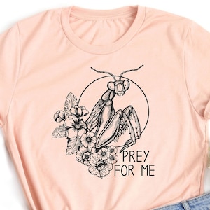 May include: A peach-colored t-shirt with a black and white illustration of a praying mantis with flowers and the text "Prey for Me".