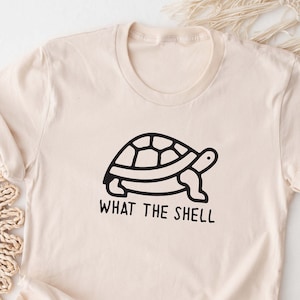 May include: A beige t-shirt with a black line drawing of a turtle and the text "What the Shell".