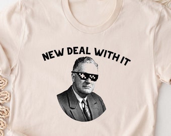 May include: Cream-colored t-shirt featuring a black and white image of a man wearing pixelated sunglasses. The text "NEW DEAL WITH IT" is arched above the image in black. The shirt has short sleeves and a crew neck.