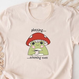 May include: A beige t-shirt with a cartoon frog sitting on a red mushroom, holding a cup of coffee. The text on the shirt reads "plotting... scheming even".