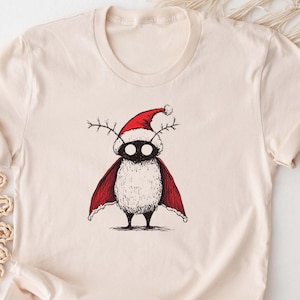 May include: A beige t-shirt with a black and white illustration of a creature wearing a red Santa hat and cape. The creature has large eyes and antlers. The text "bootees" is in the bottom left corner of the image. The text "etsy.com/shop/bootees" is in the bottom right corner of the image.