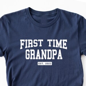 May include: Navy blue t-shirt with the text "FIRST TIME GRANDPA" in white, arched lettering. Below the text is "EST. 2023" in a rectangular box. The shirt is folded and has rolled-up sleeves.