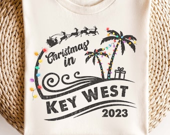 May include: A white t-shirt with a black graphic design of palm trees with Christmas lights, a wave, and the text "Christmas in Key West 2023".