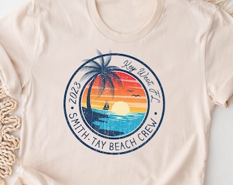 May include: A beige t-shirt with a vintage-style graphic print of a palm tree, sunset, and sailboat. The graphic is in a circle with the text "Key West FL", "2023", "Smith-Tay Beach Crew".