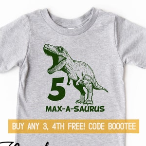 Pode incluir: Uma camiseta cinza de manga curta com um desenho de dinossauro verde, o número 5 e o texto "MAX-A-SAURUS". A camiseta tem gola redonda e é feita de um material macio. O logotipo da marca está no canto inferior esquerdo.