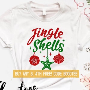 May include: White t-shirt with the words "Jingle Shells" in red and green script. The design includes red and green seashell ornaments and a starfish. The shirt also features the text "Boootees" and a list of available sizes.