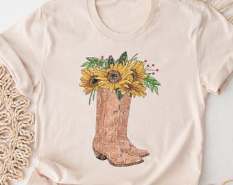 Puede incluir: Una camiseta beige con un gráfico de una bota vaquera marrón con girasoles y hojas verdes que salen de la parte superior.