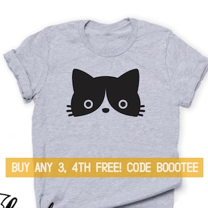 May include: A grey t-shirt with a black and white cat face graphic. The cat's face is centrally placed, with large eyes and a small nose. A yellow banner displays the text "BUY ANY 3, 4TH FREE! CODE BOOOTEE".