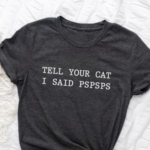 Puede incluir: Una camiseta gris oscuro con el texto "TELL YOUR CAT I SAID PSPSPS" impreso en blanco.