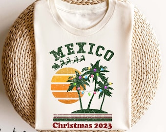 Może przedstawiać: Kremowa koszulka z postarzaną grafiką. Słowo "MEXICO" znajduje się nad zachodem słońca, palmami i saniami Świętego Mikołaja. Tekst "Christmas 2023" znajduje się poniżej. Koszulka leży na tkanej, okrągłej powierzchni.